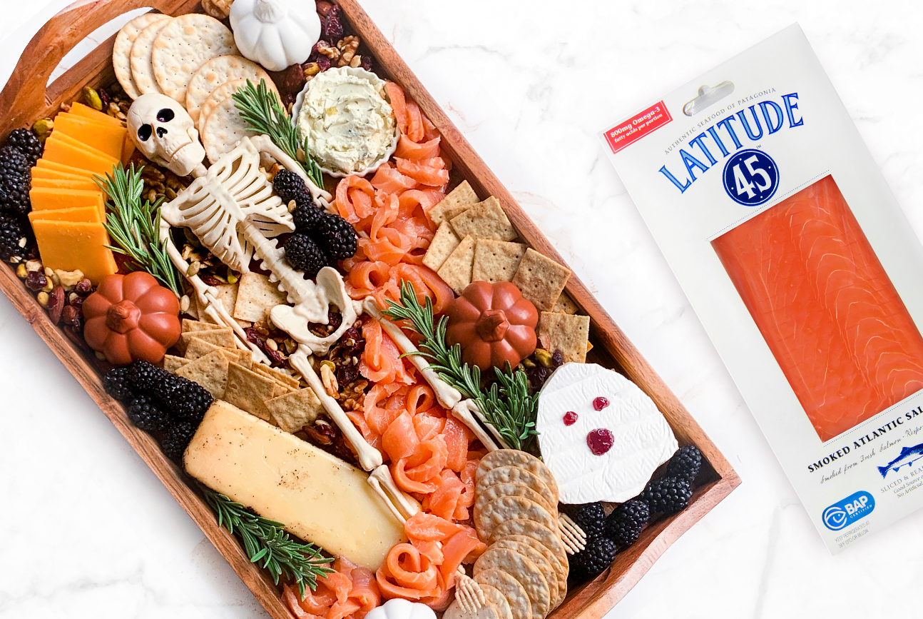a smoked salmon char-boo-terie board with a skeleton in the middle surrounded by cheeses, crackers, berries, herbs, and smoked salmon